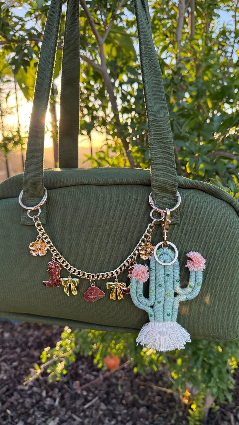 A green bag at sunset with a custom DuoAlity chain of gold bows, cowboy boots and flowers, plus a cactus keychain